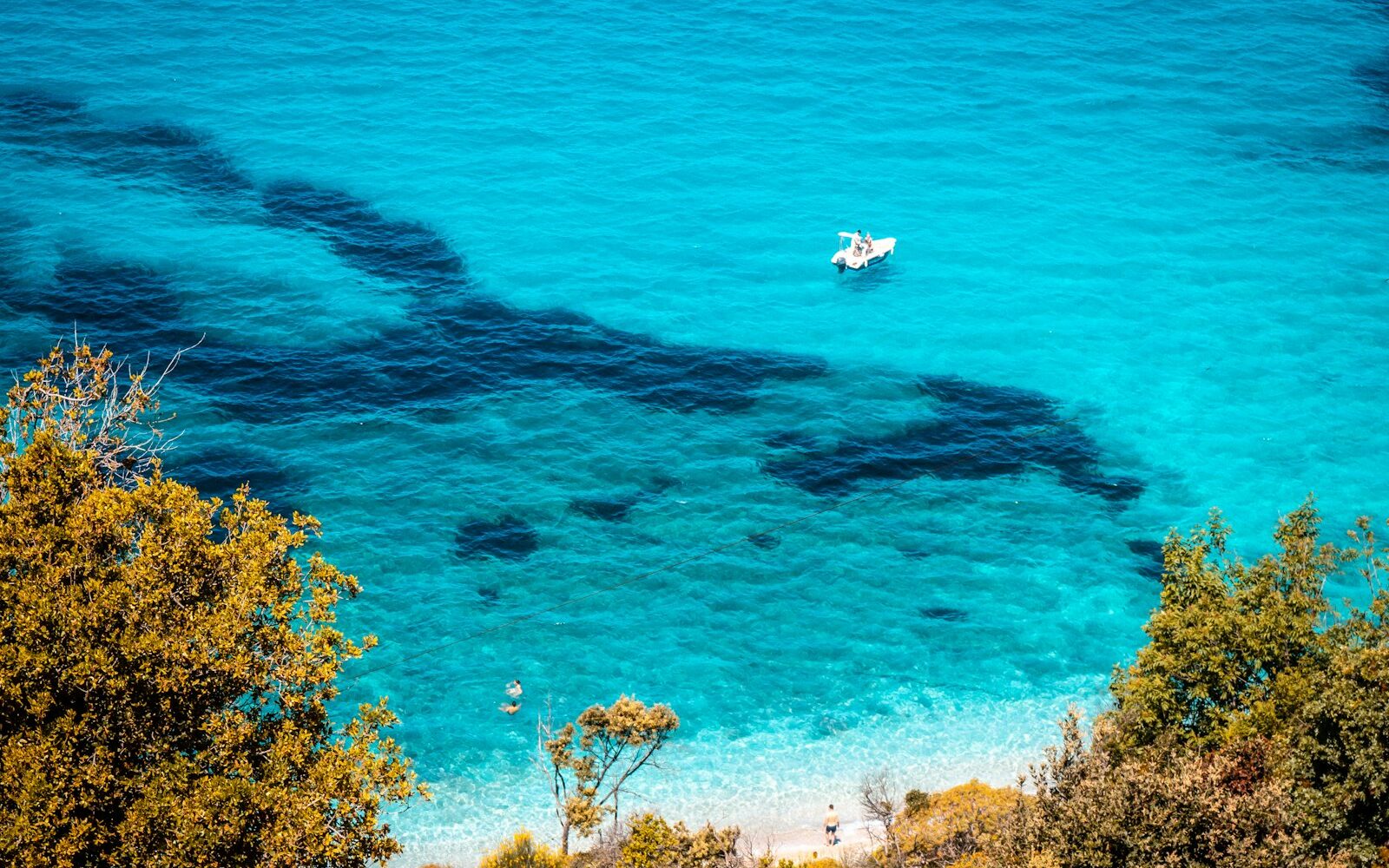 A small boat anchored in a bright turquoise cove below wooded cliffs