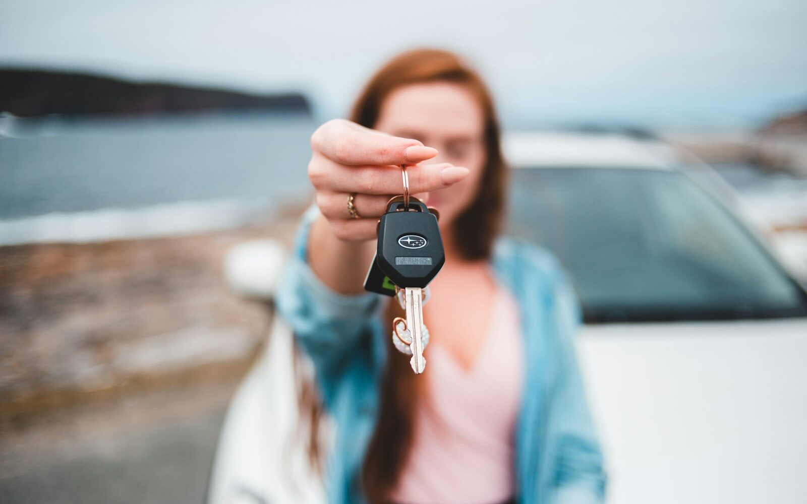 A woman holding out the keys to a white rental car, the coast behind her